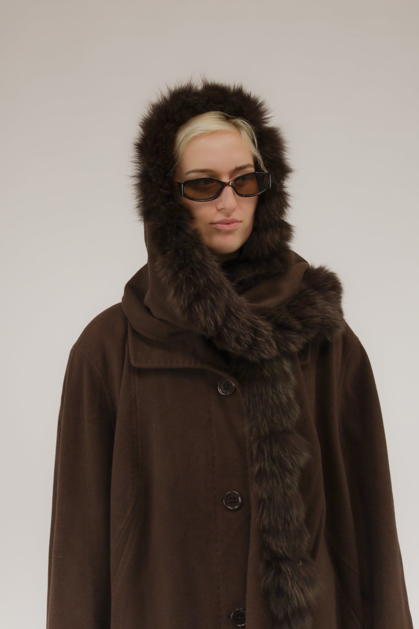 Model draagt bruine wollen mantel met afneembare kraag en capuchon met nepbont – vooraanzicht, A-lijn model.