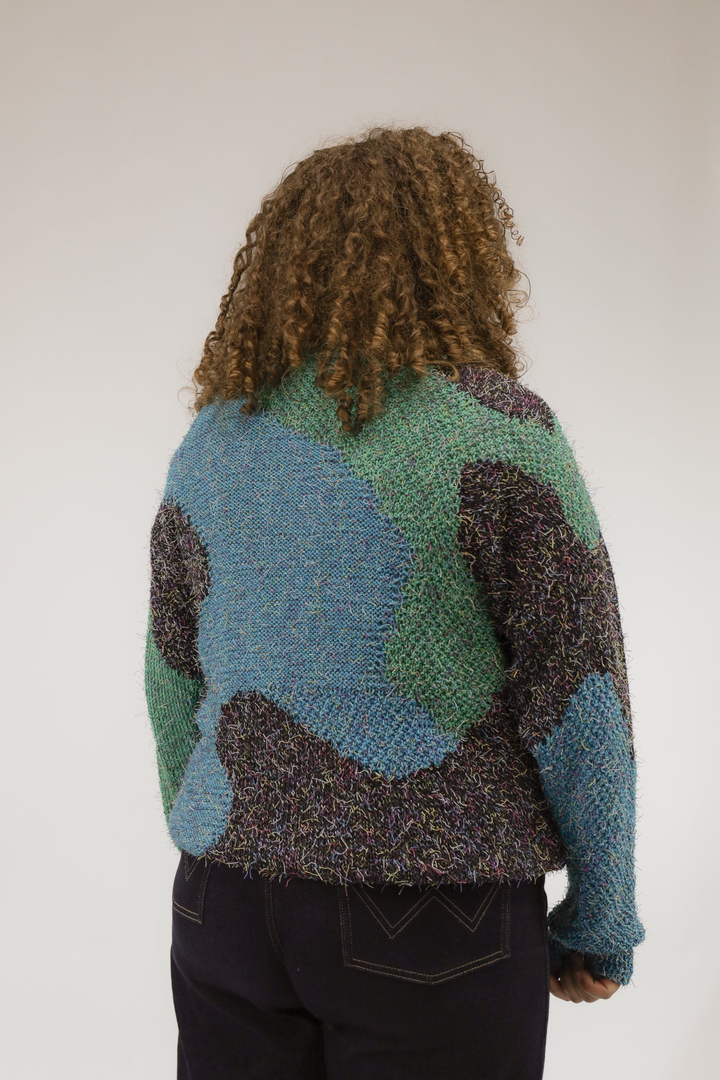 Model van achteren met vintage gebreide trui in zwart, groen en blauw – artistiek patroon en zachte textuur zichtbaar.