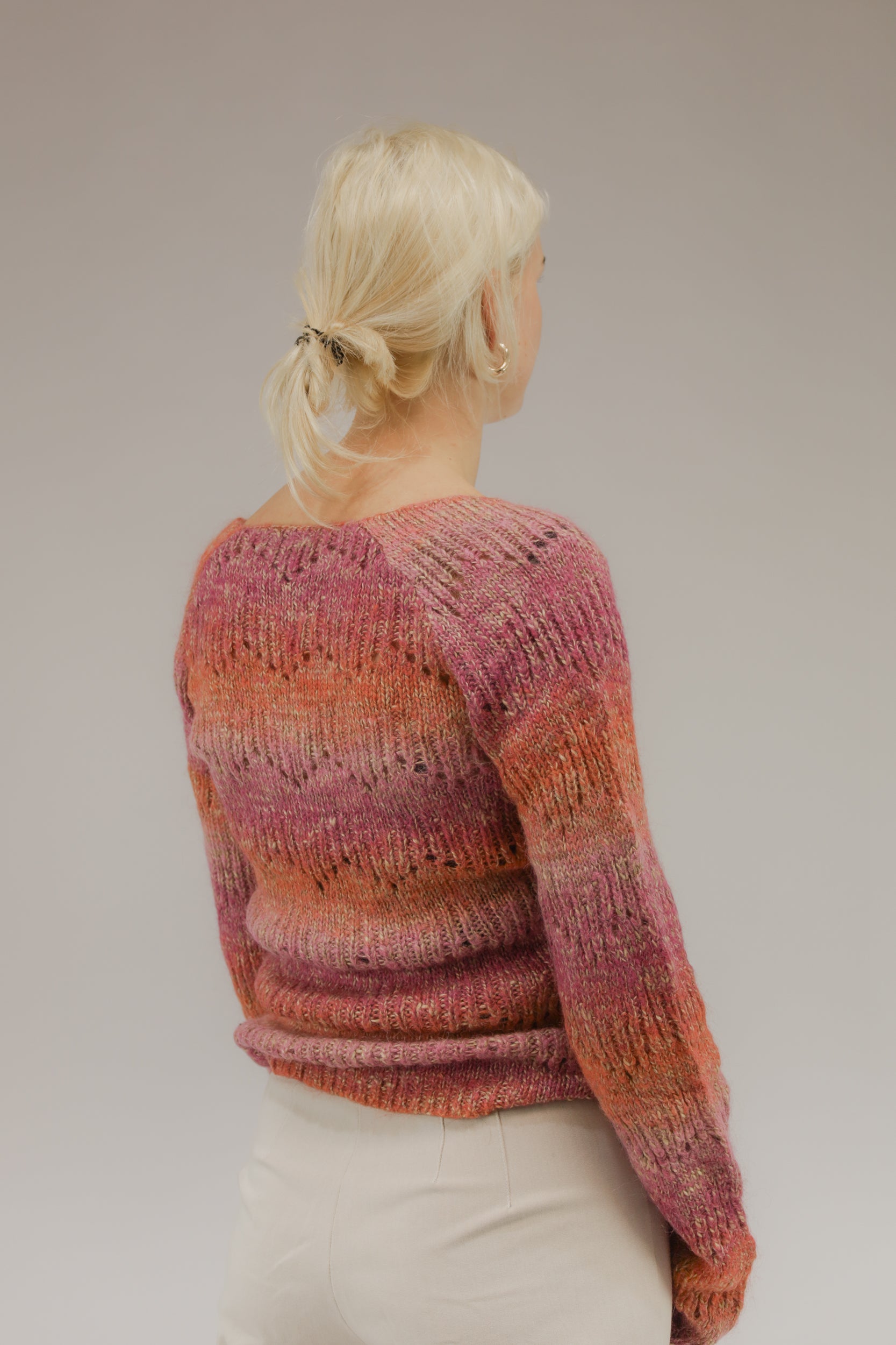 Model van achteren met fijngebreide trui in oranje, roze en beige tinten – subtiel open geweven patroon.
