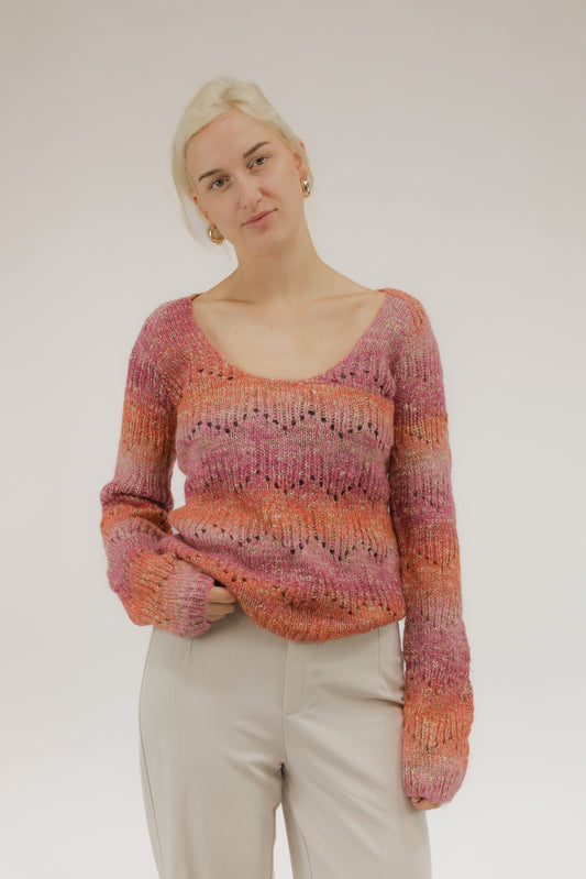 Model draagt fijngebreide trui met oranje, roze en beige streepdessin – vooraanzicht met diepe ronde hals.