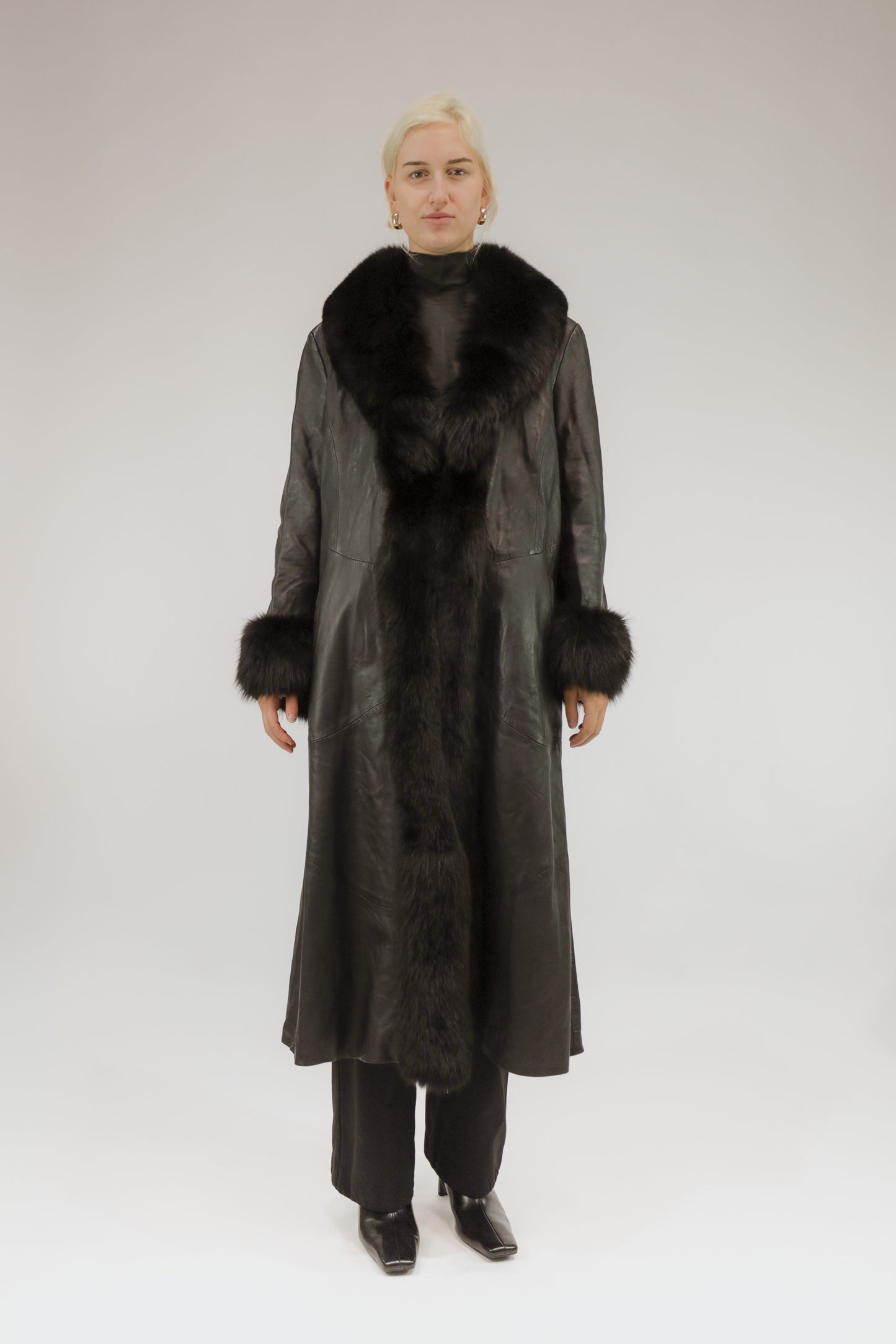 Model draagt lange zwarte vintage leren jas met bont sjaalkraag en bonten manchetten – vooraanzicht.