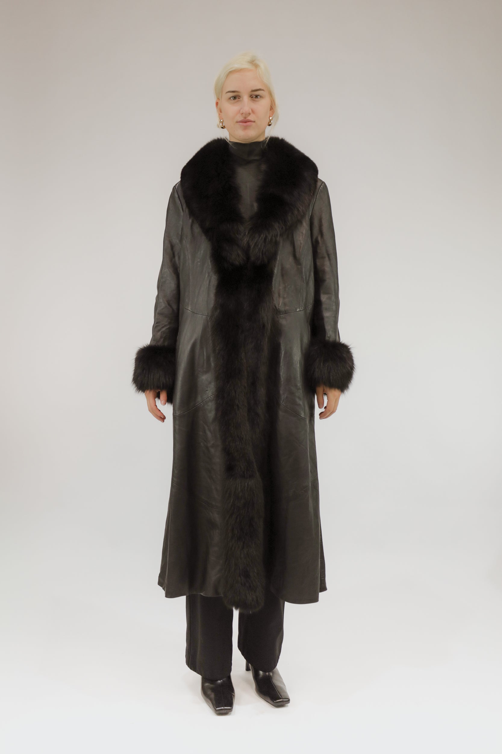 Model draagt lange zwarte vintage leren jas met bont sjaalkraag en bonten manchetten – vooraanzicht.
