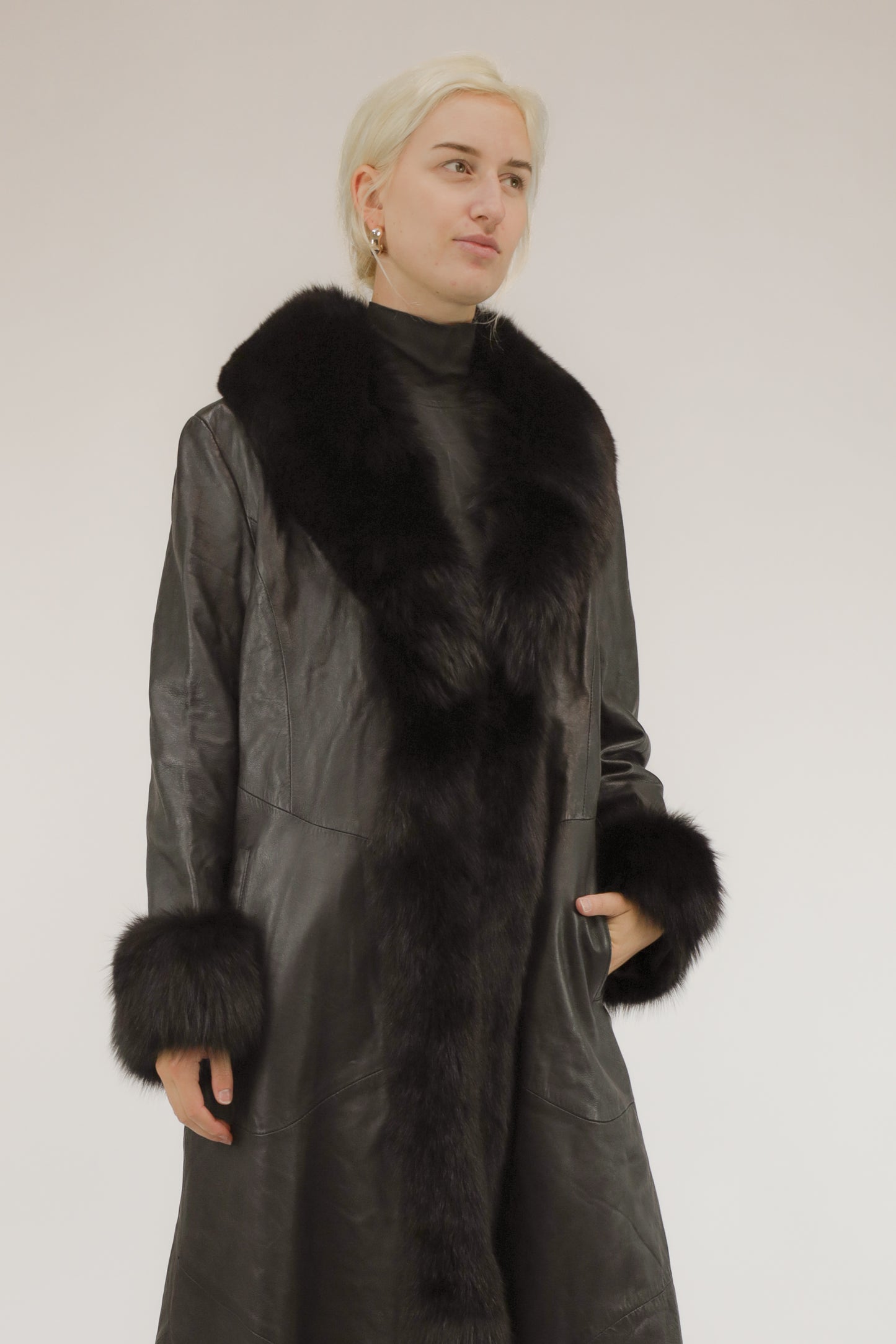 Model draagt lange zwarte vintage leren jas met bont sjaalkraag en bonten manchetten – vooraanzicht.