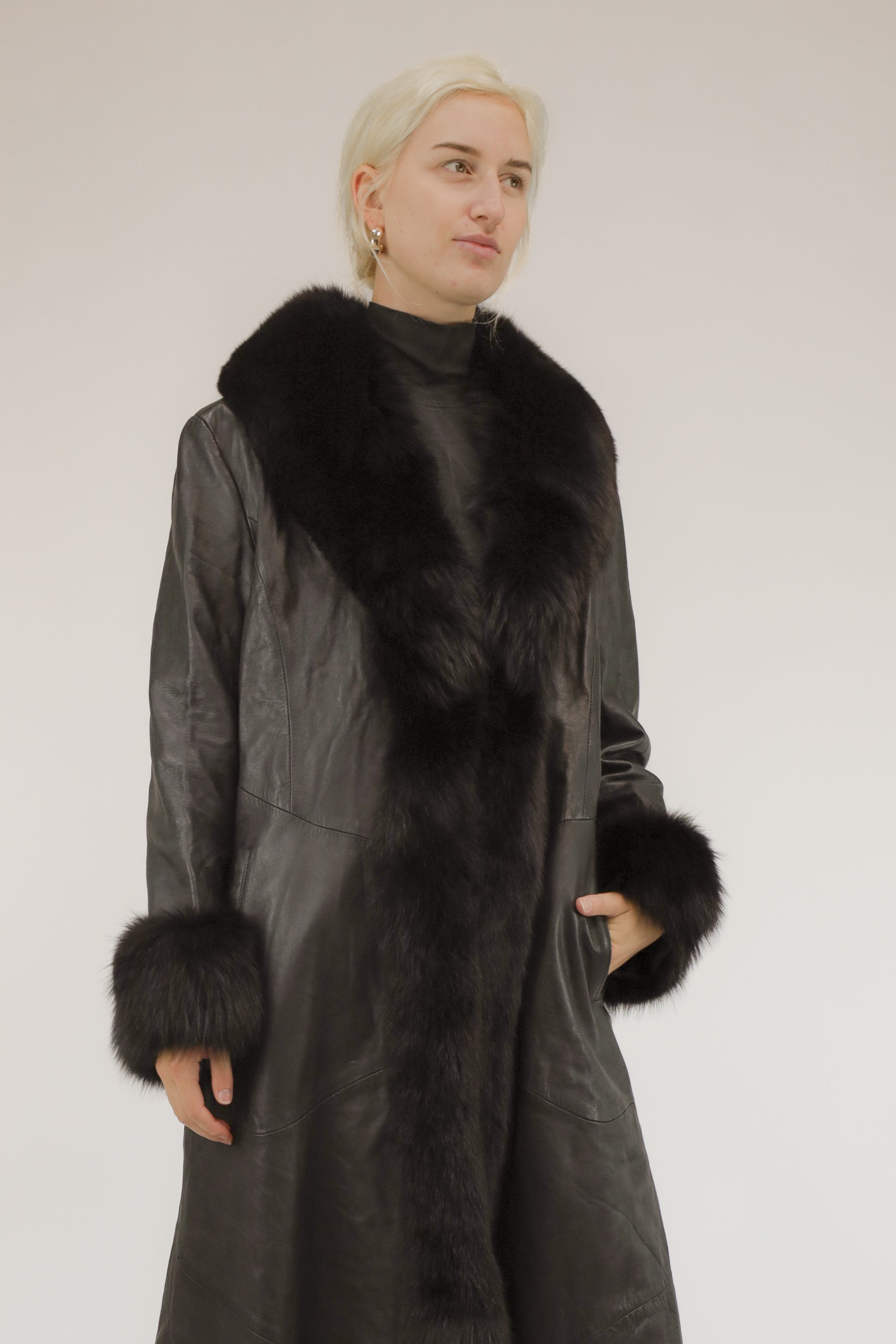 Model draagt lange zwarte vintage leren jas met bont sjaalkraag en bonten manchetten – vooraanzicht.